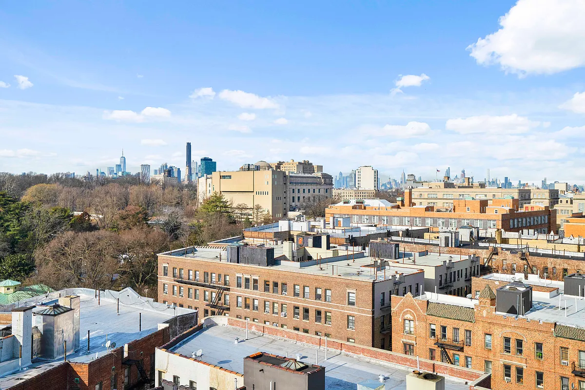 The image presents an aerial view of multiple brick apartment buildings under a clear, blue sky with scattered clouds. Several buildings exhibit flat roofs, some with visible rooftop structures and varying textures of roofing material. The cityscape extends to the horizon, showcasing distant skyscrapers and lush greenery, giving a sense of urban density blended with natural settings.