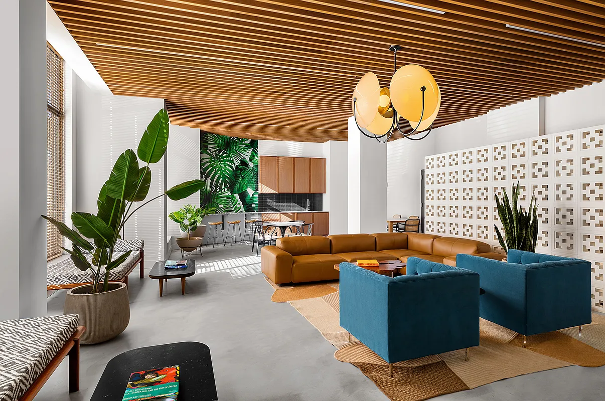 This is a stylish and modern living room with a relaxed, inviting atmosphere. A large, comfortable sectional sofa and blue armchairs anchor the space, complemented by layered area rugs on a polished concrete floor. The room features a wood slat ceiling and is decorated with large potted plants, enhancing its contemporary and natural feel.