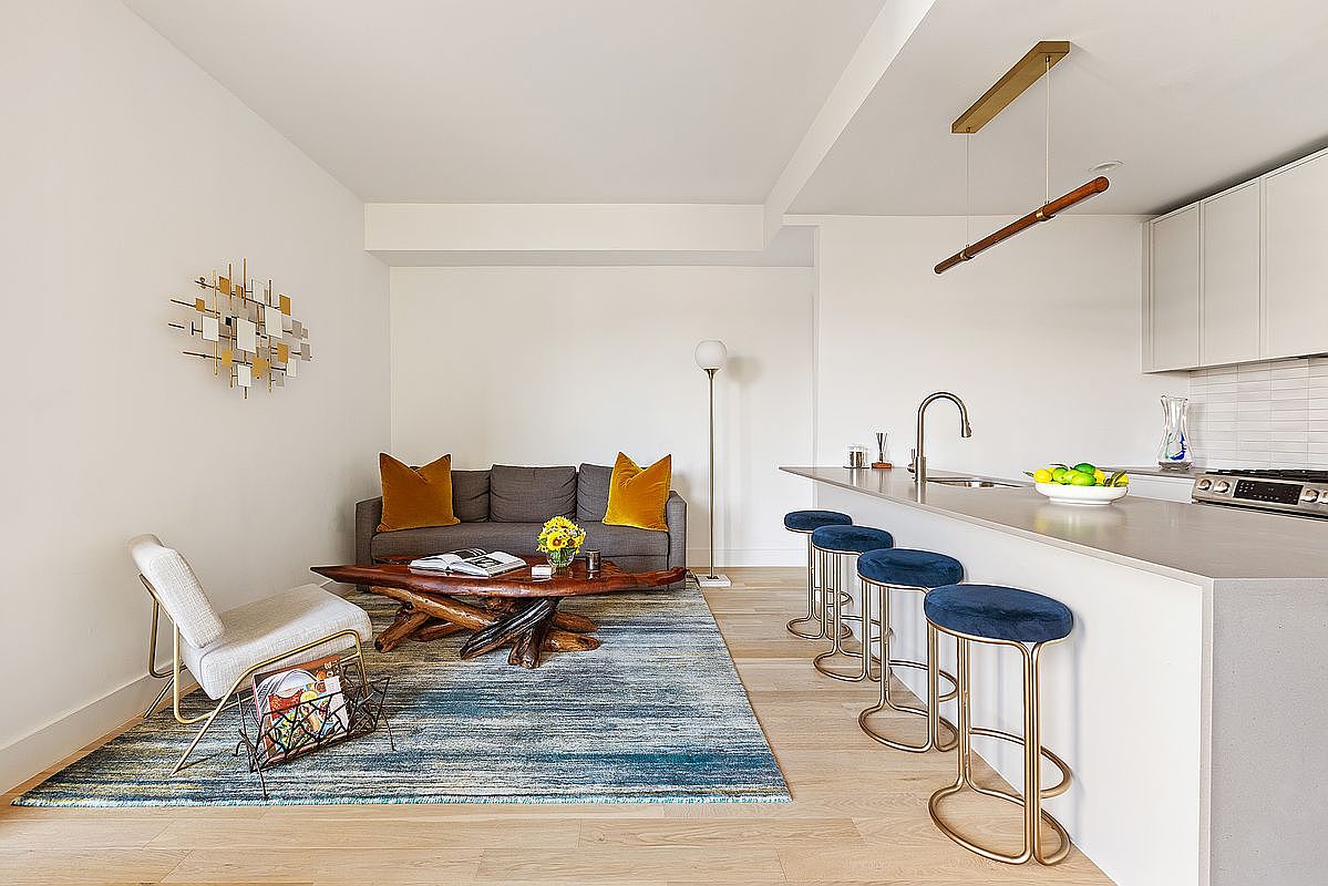 This is a well-lit, modern living room and kitchen area. The living room features a gray sofa with mustard-colored pillows, a unique wooden coffee table, and a blue and gray rug. The kitchen area has a long island with blue velvet bar stools and modern lighting fixtures, creating a stylish and inviting space.