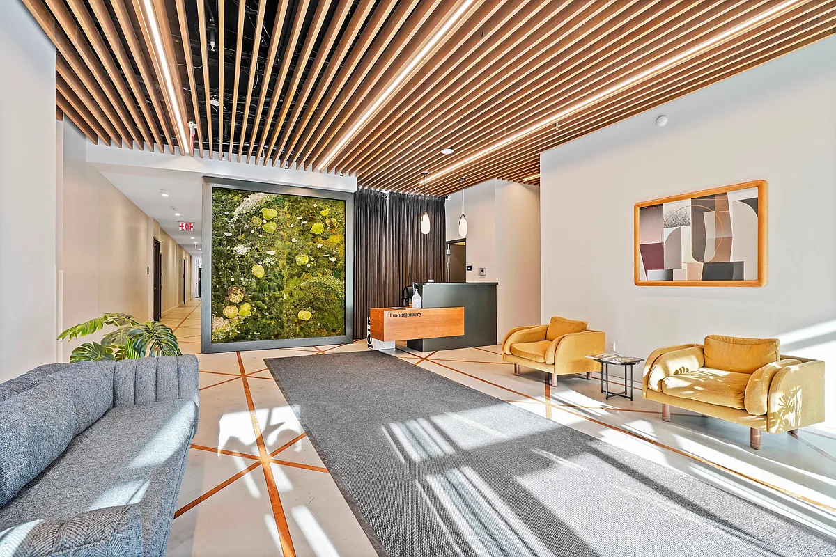 This is an interior shot of a modern building lobby. The space is characterized by a slat ceiling with integrated lighting, a large greenery wall, and contemporary furnishings including armchairs and a sofa. The flooring features a geometric pattern, enhancing the lobby's stylish and welcoming atmosphere for residents and guests.