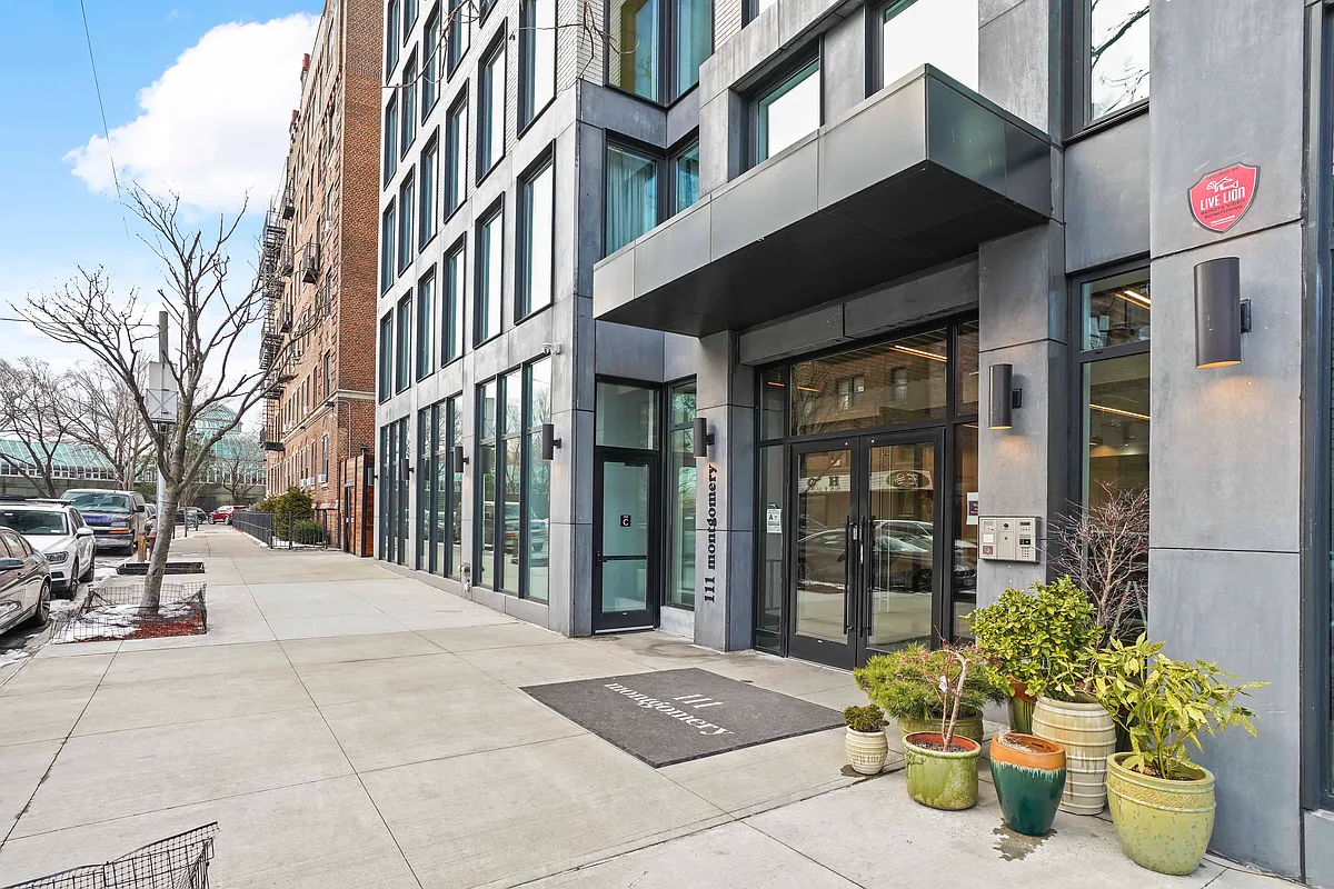 This is an exterior shot of the entrance to a modern apartment building. The building features large windows and a covered entryway with the address '111 Montgomery' displayed. Several potted plants add a touch of greenery to the entrance.