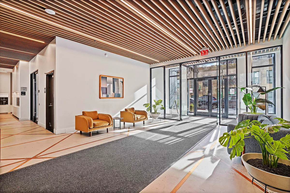 This is a modern lobby area, possibly in an apartment building or upscale office space. The design features a wooden slat ceiling with integrated lighting, contemporary furniture in mustard yellow, and potted plants creating a welcoming atmosphere. A large, gray runner guides guests towards the building's entrance, which is framed by floor-to-ceiling windows.
