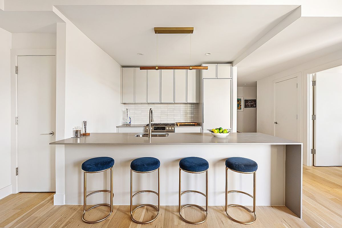 This is a bright and modern kitchen featuring a large island with seating for four, white cabinetry, and stainless steel appliances. The kitchen is well-lit with a contemporary light fixture above the island. The overall impression is clean, stylish, and functional, perfect for a modern home.