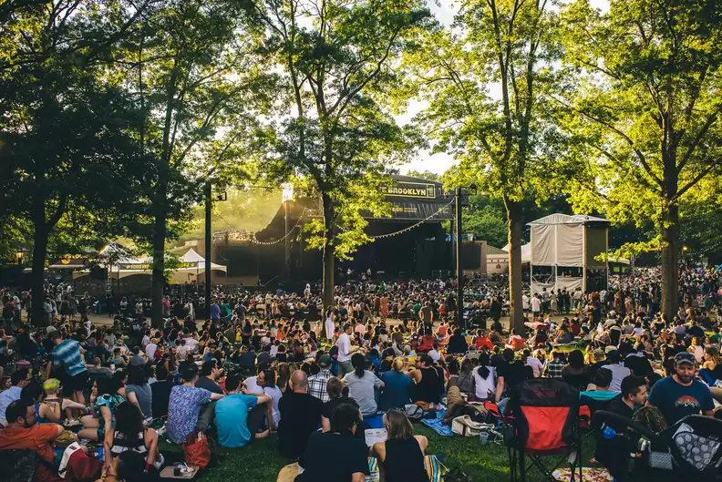 The image depicts a large outdoor event occurring in what appears to be a park or community green space. A stage is visible in the background, suggesting a concert or performance, with a large crowd of people gathered on the lawn. Tall trees surround the area, creating a natural and inviting atmosphere, enhancing the appeal of this community amenity as a recreational and social gathering space.