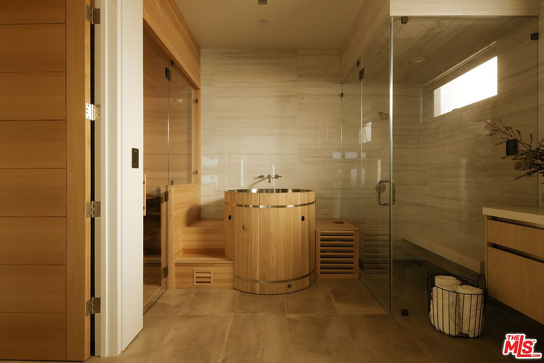 This luxurious primary bathroom features a spa-inspired design with a central wooden soaking tub, a glass-enclosed walk-in shower, and an integrated sauna. The space is finished with warm wood paneling and neutral stone tiles, creating a serene and high-end atmosphere. The perspective is a straight-on shot that highlights the seamless integration of wellness amenities within a modern residential bathroom.