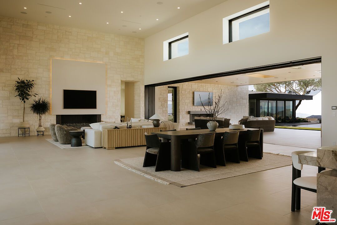 This expansive, open-concept living and dining area features soaring ceilings and a seamless transition to the outdoors through massive sliding glass walls. The space is anchored by a striking floor-to-ceiling stone fireplace wall and a sophisticated dining set, creating a luxurious and airy atmosphere. The neutral color palette and minimalist design emphasize the grand scale and high-end architectural details of this modern residence.