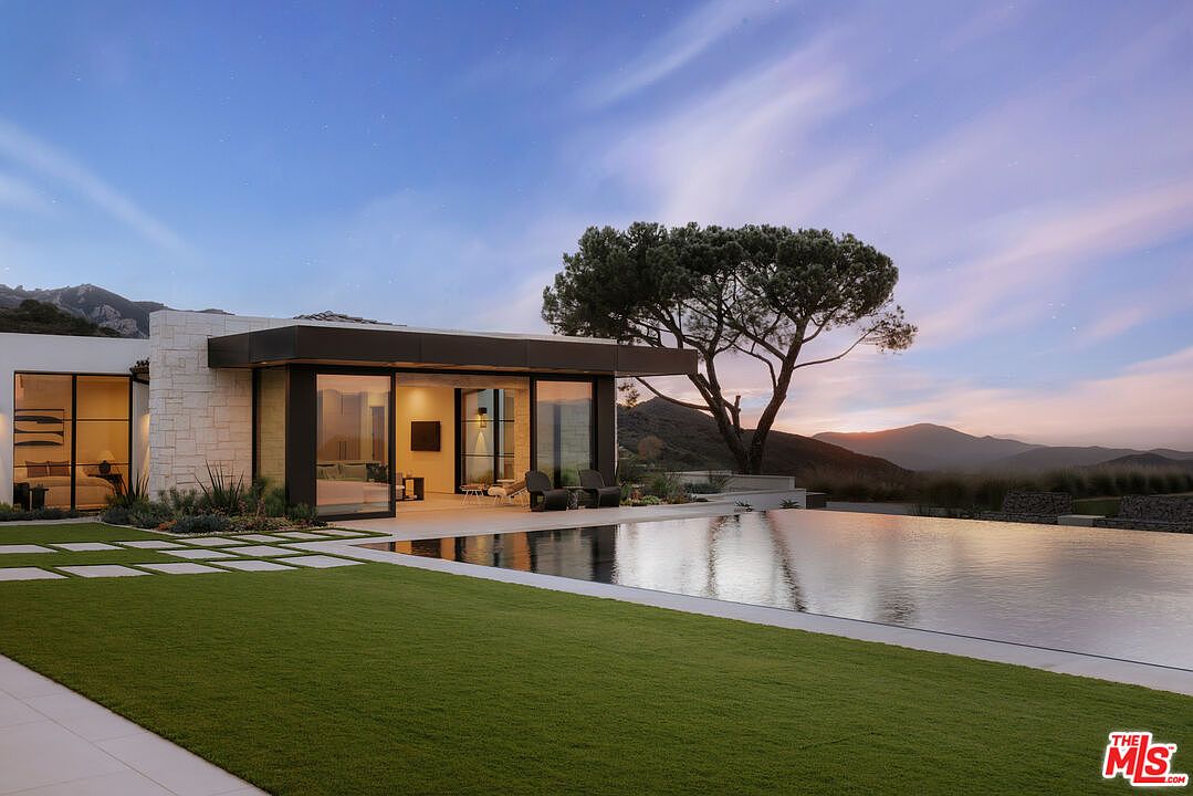 This stunning modern residence features a seamless indoor-outdoor flow, highlighted by a sleek infinity-edge pool that reflects the twilight sky. The architecture emphasizes clean lines, expansive glass walls, and natural stone textures, set against a backdrop of rolling hills and a mature tree. The composition captures a serene, luxurious atmosphere perfect for high-end real estate marketing.