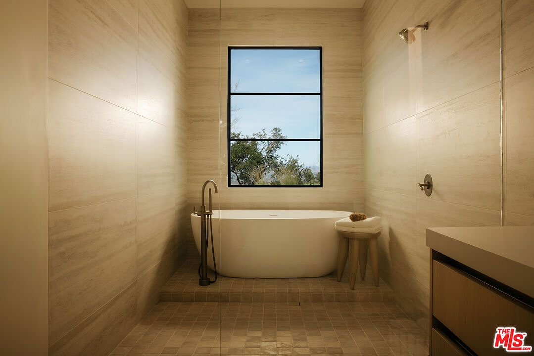 This luxurious primary bathroom features a sleek, freestanding soaking tub positioned on a raised platform against a large window with a scenic view. The space is finished with elegant, light-toned stone tile walls and a matching tiled floor, creating a serene and spa-like atmosphere. A minimalist wooden stool with towels sits beside the tub, enhancing the clean, contemporary aesthetic of the room.