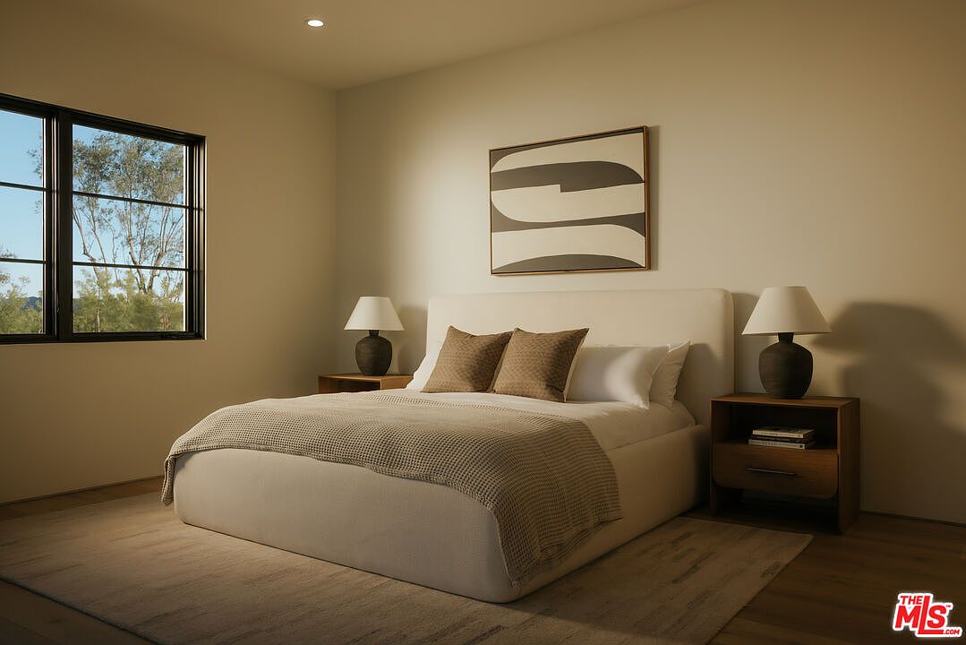 This serene primary bedroom features a plush, upholstered bed frame centered against a neutral-toned wall adorned with minimalist abstract art. The room is flanked by warm wooden nightstands topped with matching table lamps, creating a balanced and inviting atmosphere. A large window to the left allows soft natural light to filter in, highlighting the clean lines and contemporary aesthetic of the space.