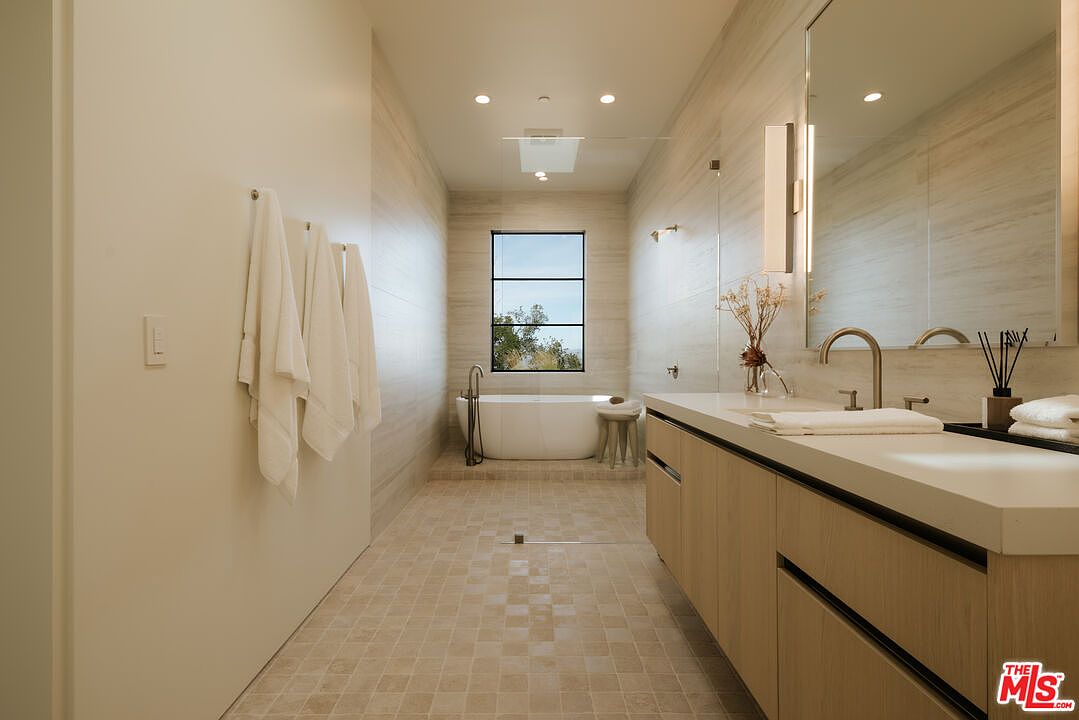 This luxurious primary bathroom features a sleek, modern design with light wood cabinetry, a double vanity, and a freestanding soaking tub positioned beneath a large window. The space is finished with neutral, textured wall tiles and a grid-patterned floor, creating a serene and spa-like atmosphere. The perspective is a wide-angle shot from the entrance, showcasing the clean lines and high-end finishes of the room.