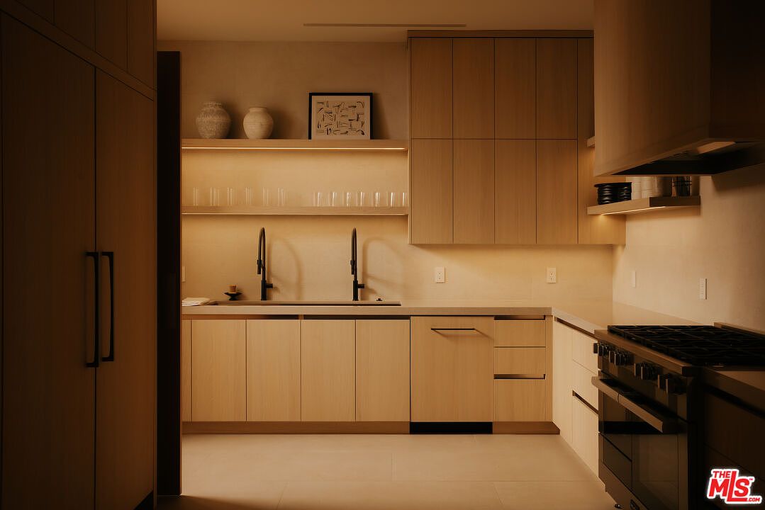 This modern, minimalist kitchen features light wood cabinetry, sleek integrated appliances, and warm under-cabinet lighting that creates a sophisticated, inviting atmosphere. The space is anchored by a clean countertop with dual faucets and open shelving adorned with decorative pottery and glassware. The overall aesthetic is contemporary and refined, emphasizing clean lines and a warm, monochromatic color palette.