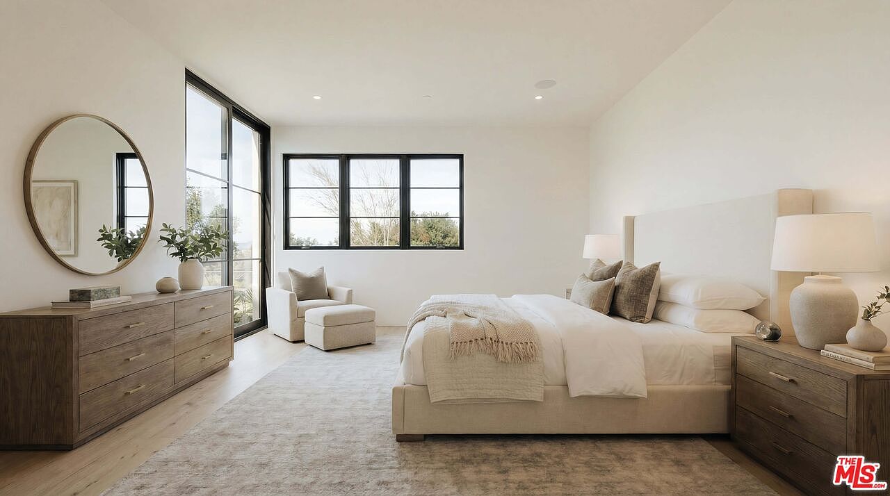 This spacious and serene primary bedroom features a neutral color palette, a plush upholstered bed, and a matching wooden dresser. Large windows and a glass door allow for abundant natural light, creating an airy and inviting atmosphere. The room is accented by a round mirror, a cozy armchair with an ottoman, and elegant bedside lamps, reflecting a sophisticated, modern aesthetic.