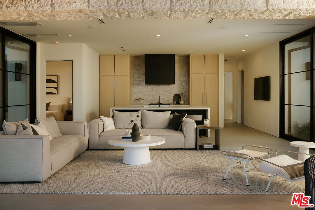 This sophisticated open-concept living area features a neutral color palette, plush contemporary sofas, and a minimalist round coffee table centered on a textured area rug. The space is anchored by a sleek kitchen island in the background, framed by light wood cabinetry and a textured stone wall, creating a seamless blend of comfort and modern luxury. The perspective is a wide, eye-level shot that emphasizes the airy, high-end design and the cohesive flow between the living and kitchen zones.