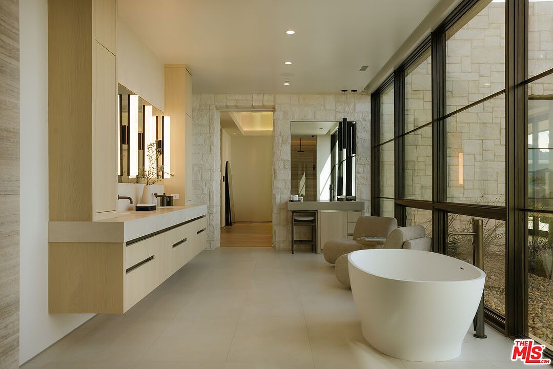 This luxurious primary bathroom features a sleek, minimalist design with a floating light-wood vanity, a freestanding soaking tub, and floor-to-ceiling windows that offer a serene view of the outdoors. The space is anchored by textured stone walls and large-format floor tiles, creating a sophisticated, spa-like atmosphere. The perspective is wide and inviting, showcasing the seamless blend of modern architecture and natural light.
