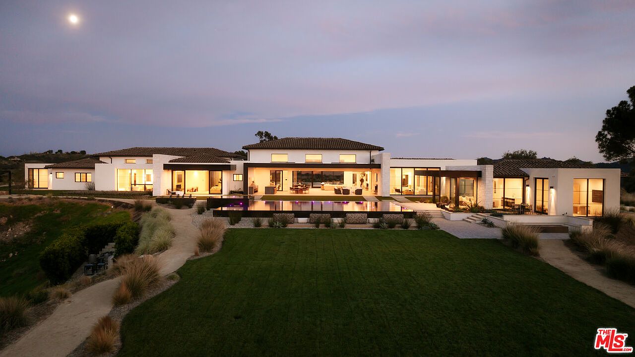 This stunning contemporary estate is captured at twilight, showcasing a sprawling rear facade with floor-to-ceiling glass walls that illuminate the interior. The property features a sleek infinity-edge pool, a manicured lawn, and a sophisticated architectural design that seamlessly blends indoor and outdoor living spaces. The wide-angle perspective highlights the expansive layout and the serene, elevated setting against a soft evening sky.