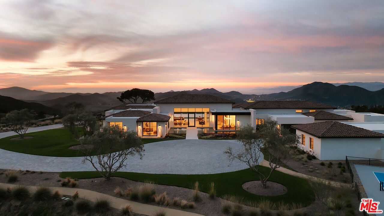 This stunning aerial shot captures a sprawling, modern luxury estate set against a dramatic, colorful sunset over rolling mountains. The home features a contemporary architectural design with a large circular driveway, manicured lawns, and warm interior lighting glowing through expansive glass windows. The perspective emphasizes the property's grand scale and its serene, elevated location, creating an inviting and sophisticated atmosphere.