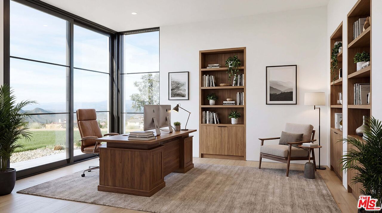 This sophisticated home office features a warm, modern aesthetic with rich wood furniture, including a substantial executive desk and built-in shelving units. Large floor-to-ceiling windows flood the room with natural light and provide a scenic view of the surrounding landscape, while a plush area rug and comfortable armchair create an inviting atmosphere. The clean lines and minimalist decor contribute to a professional yet serene workspace.