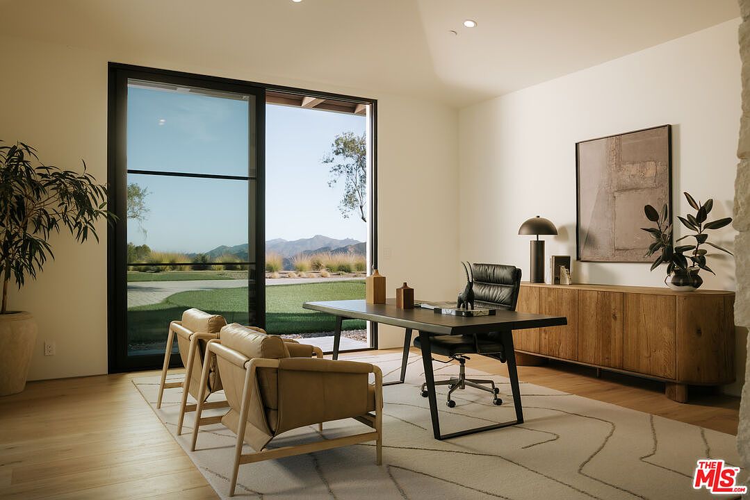 This sophisticated home office features a minimalist design with a sleek dark wood desk, a black leather executive chair, and two tan leather guest chairs. A large sliding glass door provides an expansive, unobstructed view of the rolling hills and manicured landscape outside, bathing the room in natural light. The space is accented by a warm wooden credenza, abstract wall art, and a potted plant, creating a professional yet serene atmosphere.