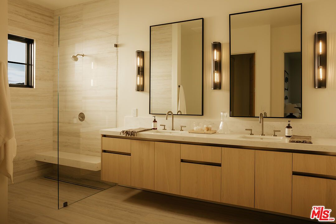 This modern primary bathroom features a sleek, floating light-wood vanity with dual sinks and a clean white countertop. The space is accented by two rectangular mirrors, industrial-style wall sconces, and a spacious walk-in shower with a glass partition and neutral stone tiling. The overall aesthetic is minimalist and sophisticated, creating a bright and serene atmosphere.