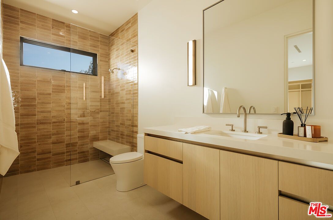 This modern, minimalist bathroom features a sleek floating vanity with light wood cabinetry and a clean white countertop. A spacious walk-in shower with textured, earth-toned tile walls and a built-in bench is enclosed by a frameless glass panel, while a large, frameless mirror reflects the bright, airy space. The room is illuminated by a contemporary vertical wall sconce, creating a sophisticated and serene atmosphere.
