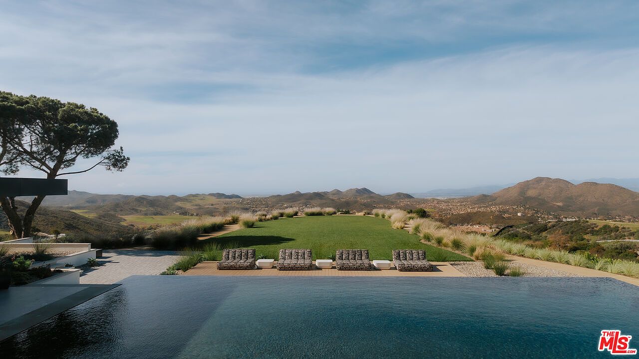 This stunning infinity-edge pool overlooks a vast, rolling landscape of hills and valleys, creating a seamless visual connection between the water and the horizon. A manicured lawn sits between the pool deck and the natural terrain, accented by ornamental grasses and a row of modern lounge chairs. The perspective is captured from a slightly elevated vantage point, emphasizing the luxurious, serene atmosphere of this high-end outdoor living space.