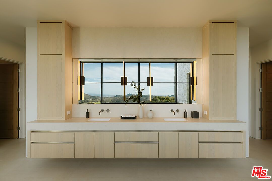 This modern primary bathroom features a sleek, floating double vanity with light wood cabinetry and a clean, white countertop. A large, horizontal window serves as the focal point, offering a panoramic view of the surrounding hills and landscape. The minimalist design is complemented by integrated lighting and high-end fixtures, creating a serene and luxurious atmosphere.