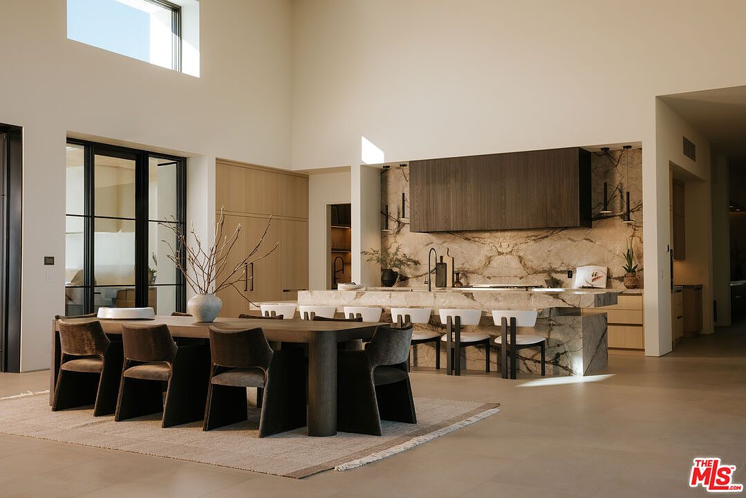 This sophisticated open-concept space seamlessly blends a formal dining area with a high-end, modern kitchen. The kitchen features a striking marble waterfall island, dark wood cabinetry, and integrated appliances, all set against a dramatic marble backsplash. The high ceilings and minimalist aesthetic create an airy, luxurious atmosphere perfect for both entertaining and daily living.