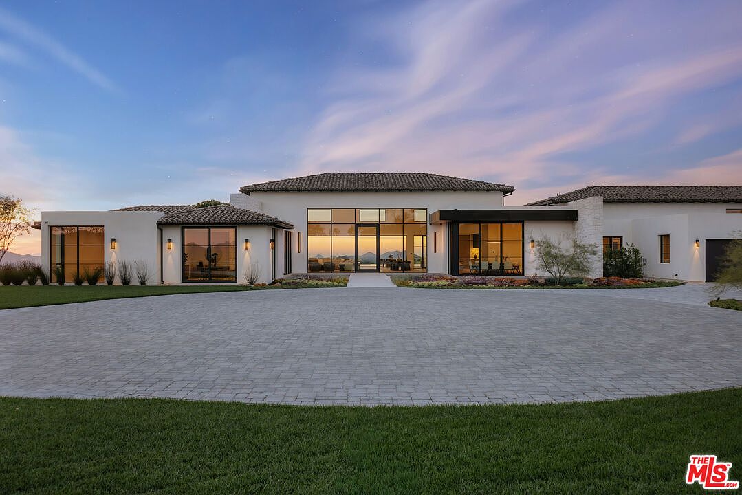 This stunning contemporary estate features a clean, minimalist architectural design with a sprawling paved circular driveway leading to a grand glass-paneled entrance. The exterior is finished in crisp white stucco with dark-framed windows and a classic tiled roof, set against a backdrop of a serene, twilight sky. The wide-angle perspective emphasizes the home's impressive scale, symmetry, and seamless integration with the surrounding landscape.