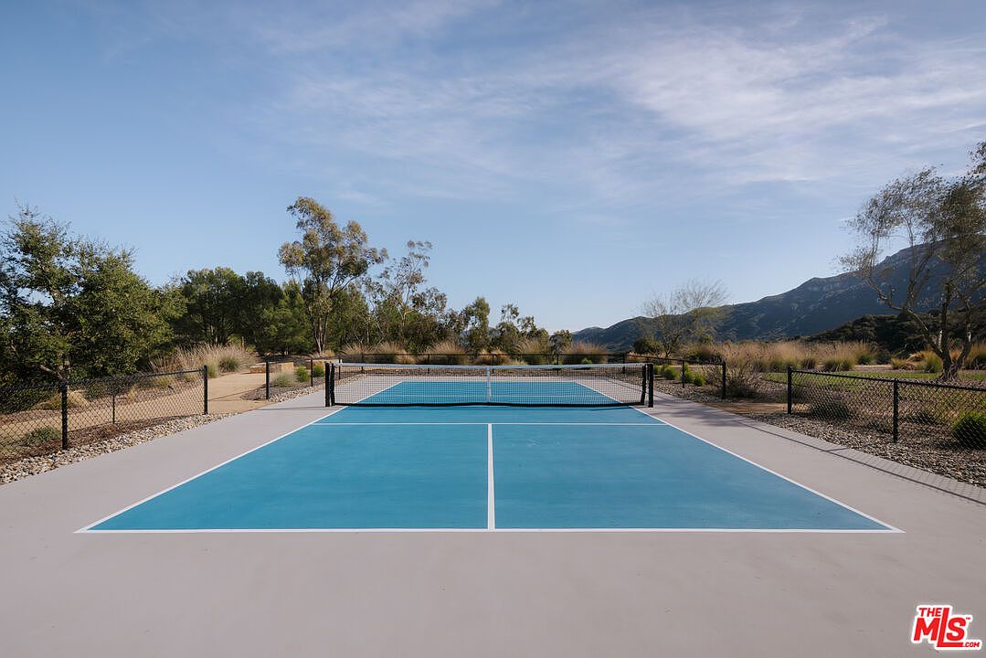 This image showcases a pristine, outdoor pickleball court set against a backdrop of rolling hills and a clear blue sky. The court features a vibrant blue playing surface with crisp white lines, enclosed by a black chain-link fence and surrounded by natural, arid landscaping. The perspective is centered and eye-level, emphasizing the recreational appeal and the expansive, scenic outdoor environment.