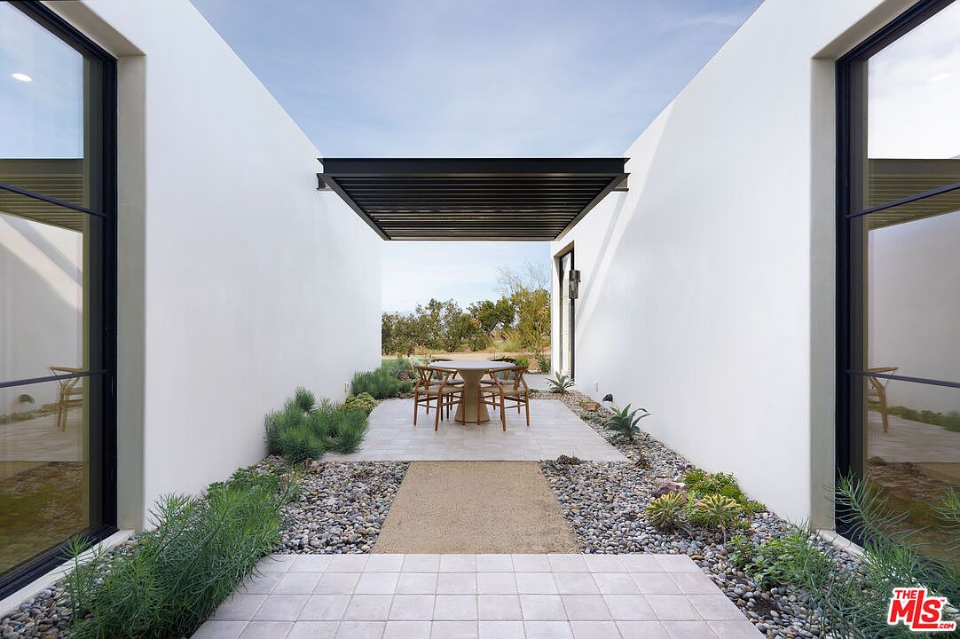This modern, minimalist outdoor patio features a central dining area sheltered by a sleek, black slatted pergola. The space is framed by two white walls and large glass windows, with a pathway of light-colored pavers surrounded by decorative gravel and desert-inspired landscaping. The perspective is centered and symmetrical, creating a serene and sophisticated transition between the indoor and outdoor living spaces.