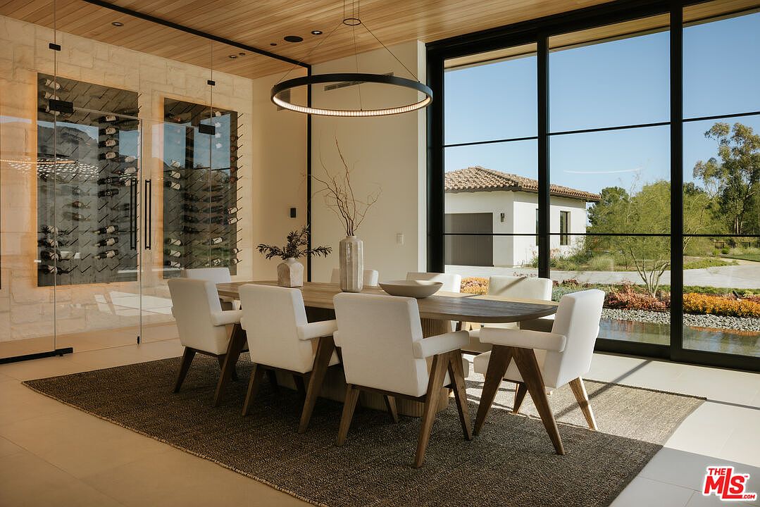 This sophisticated dining room features a minimalist, modern aesthetic with a large wooden table surrounded by six white upholstered chairs. A striking circular pendant light hangs above the table, while a glass-enclosed wine cellar wall adds a touch of luxury to the space. Large floor-to-ceiling windows provide a seamless connection to the outdoors, bathing the room in natural light and offering a serene view of the landscape.
