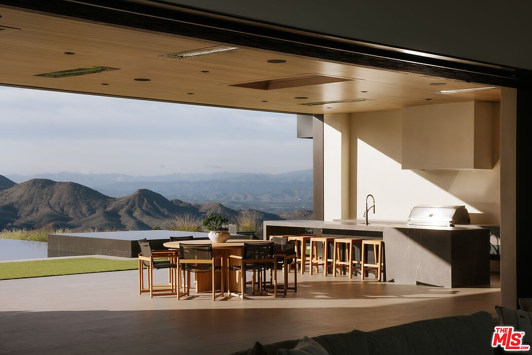 This stunning indoor-outdoor living space features a seamless transition from a modern kitchen area to a spacious covered patio overlooking a breathtaking mountain landscape. The area is furnished with a circular wooden dining table and a sleek kitchen island with bar seating, all set against a backdrop of an infinity-edge pool and expansive desert vistas. The architectural design emphasizes clean lines, natural materials, and an open-concept layout that maximizes the panoramic views.