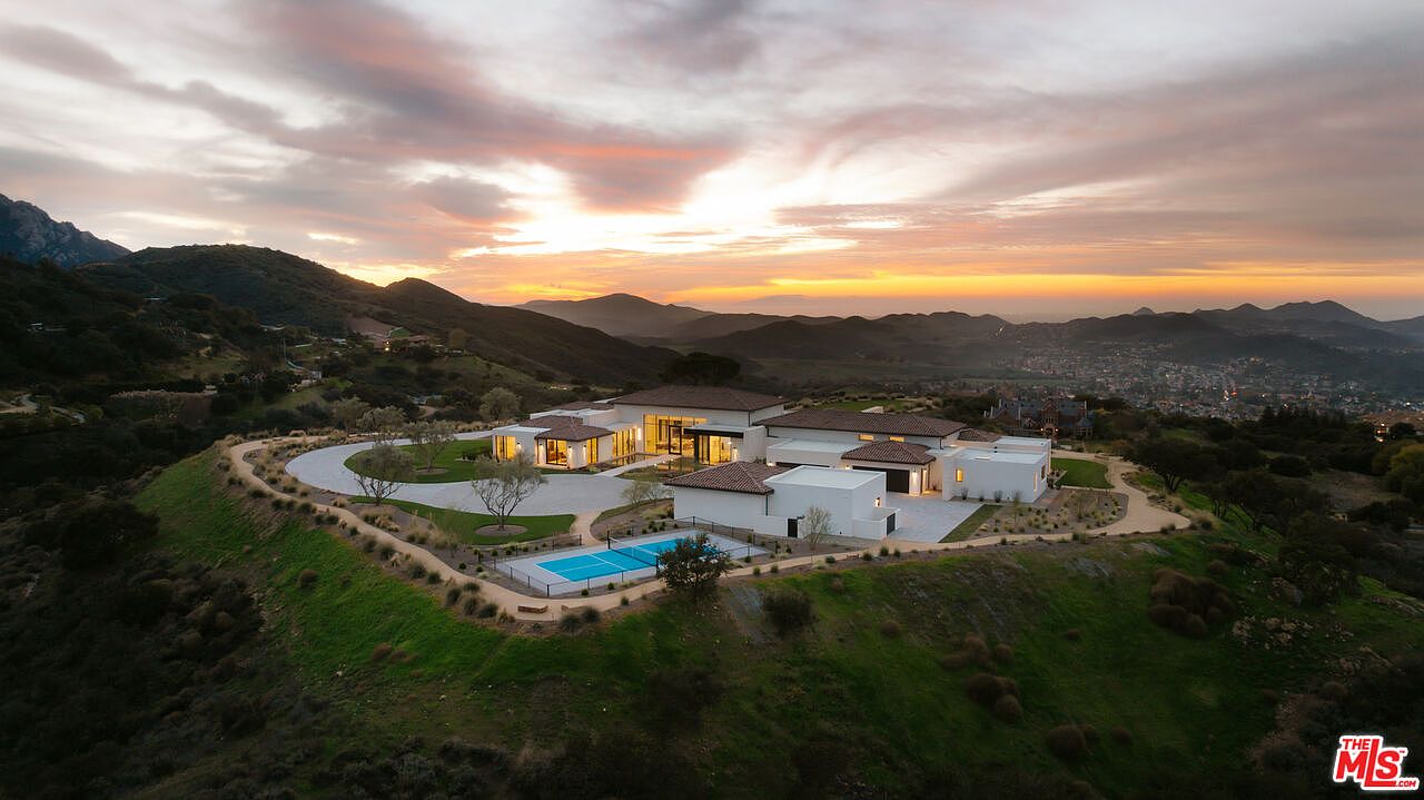 This stunning aerial view captures a sprawling, modern luxury estate perched atop a hillside at sunset. The property features a contemporary architectural design with white stucco walls, a multi-section roofline, and a private swimming pool, all set against a dramatic backdrop of rolling mountains and a distant valley town. The wide-angle perspective emphasizes the home's secluded, elevated position and its seamless integration with the expansive natural landscape.