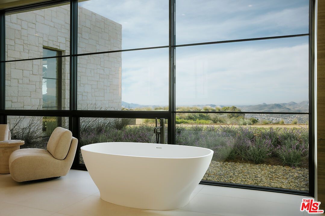 This luxurious primary bathroom features a sleek, freestanding white soaking tub positioned in front of a massive floor-to-ceiling window that frames a stunning panoramic mountain view. To the left, a modern, plush armchair sits on a light-colored floor, creating a serene and sophisticated relaxation space. The minimalist design emphasizes clean lines and a seamless connection between the indoor sanctuary and the natural landscape outside.