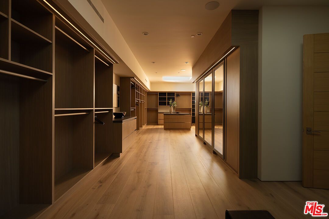 This expansive, high-end walk-in closet features custom warm-toned wood cabinetry, integrated LED lighting, and a central island with storage drawers. The space is finished with light hardwood flooring and mirrored wardrobe doors, creating a sophisticated and organized atmosphere. The perspective is a centered, eye-level shot that emphasizes the depth and luxurious scale of the dressing area.