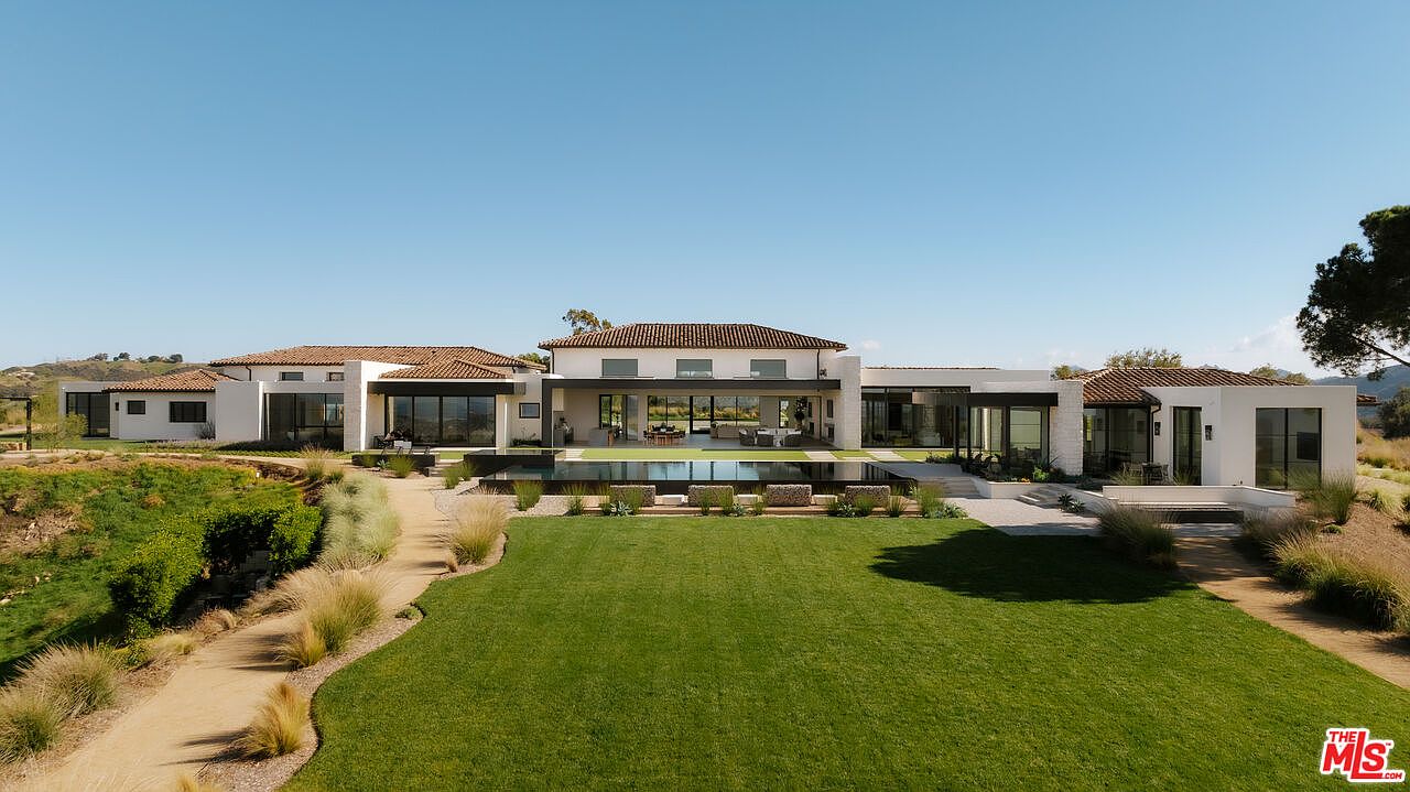This expansive, modern luxury estate features a sprawling single-story layout with a classic Mediterranean-inspired tile roof and clean, white stucco walls. The property boasts a large, manicured lawn leading to a sleek infinity-edge pool that perfectly frames the home's open-concept living area. The perspective is a wide, eye-level shot that emphasizes the seamless integration between the indoor living spaces and the vast, sun-drenched outdoor landscape.