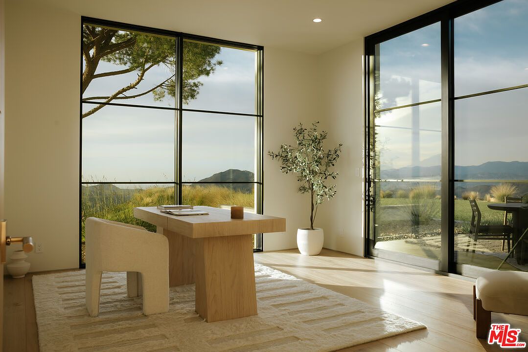 This serene home office features a minimalist aesthetic with a light wood desk, a textured cream-colored chair, and a plush area rug. Floor-to-ceiling black-framed windows flood the room with natural light and offer expansive, picturesque views of the surrounding hills and landscape. A potted olive tree adds a touch of organic greenery, creating a calm and productive atmosphere.