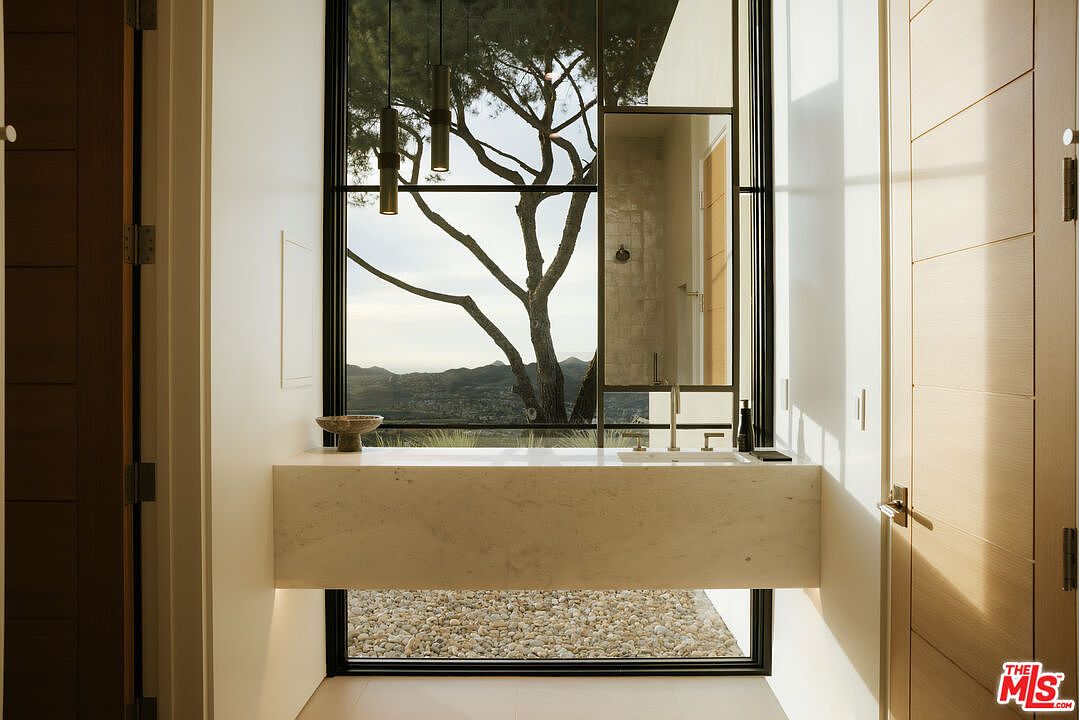 This sophisticated primary bathroom features a floating marble vanity set against a floor-to-ceiling window that frames a stunning, serene landscape view. The minimalist design is complemented by sleek, modern fixtures and warm wood-toned doors, creating a tranquil and luxurious atmosphere. The perspective is centered on the vanity, emphasizing the seamless integration of indoor luxury and the natural environment.
