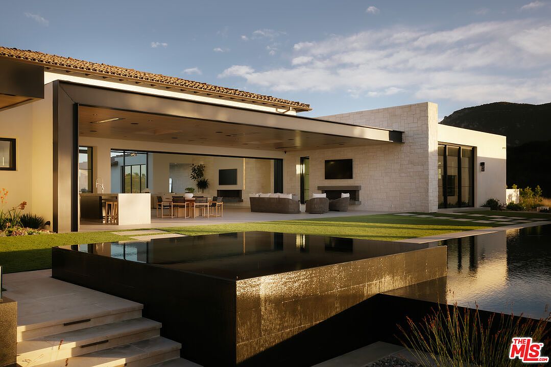 This stunning contemporary estate features a seamless indoor-outdoor living space, highlighted by a massive covered patio that opens directly into the main living area. In the foreground, a sleek, dark-tiled infinity-edge pool creates a dramatic reflection of the modern architecture and surrounding landscape. The combination of clean lines, stone accents, and expansive glass walls offers a sophisticated, resort-like atmosphere perfect for luxury entertaining.