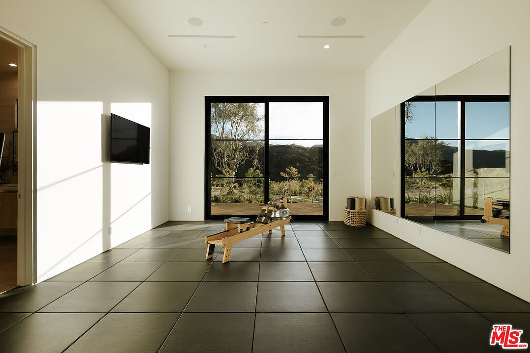 This minimalist home gym features sleek black floor tiles and a large wall-to-wall mirror that enhances the sense of space. A wooden rowing machine sits centrally in the room, positioned to face a large glass sliding door that offers a serene view of the natural landscape outside. The room is bright and airy, with a wall-mounted television and recessed lighting, creating a clean and focused environment for exercise.