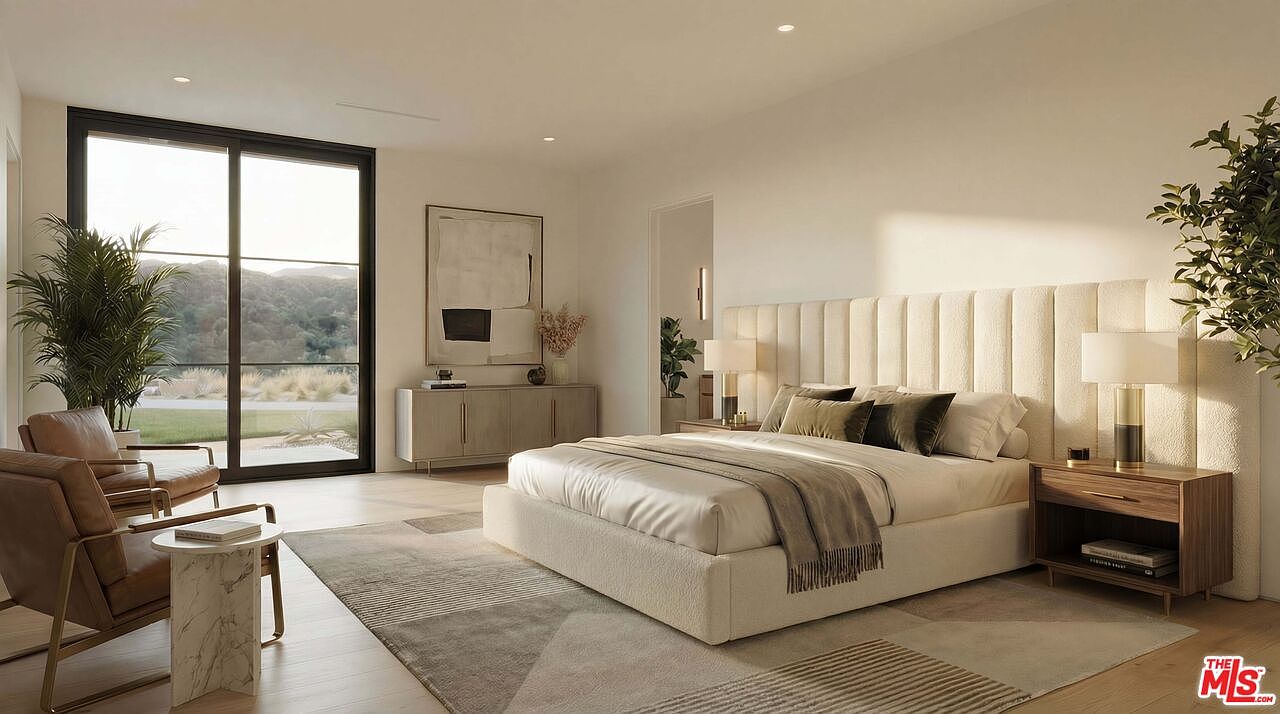 This sophisticated primary bedroom features a neutral color palette, a plush upholstered headboard, and a large area rug that anchors the space. A pair of leather armchairs sits near a floor-to-ceiling window, providing a serene view of the outdoors, while a sleek console table and matching nightstands complete the modern, minimalist aesthetic. The room is bathed in warm, natural light, creating an inviting and tranquil atmosphere.