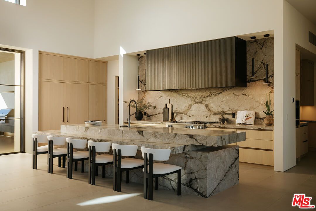 This sophisticated, high-end kitchen features a massive, waterfall-edge marble island that serves as the centerpiece, complemented by five modern, minimalist bar stools. The space showcases a striking contrast between the light-toned wood cabinetry and the dramatic, dark-veined marble backsplash and range hood. The perspective is a wide, eye-level shot that emphasizes the open-concept layout, clean lines, and luxurious, contemporary aesthetic.