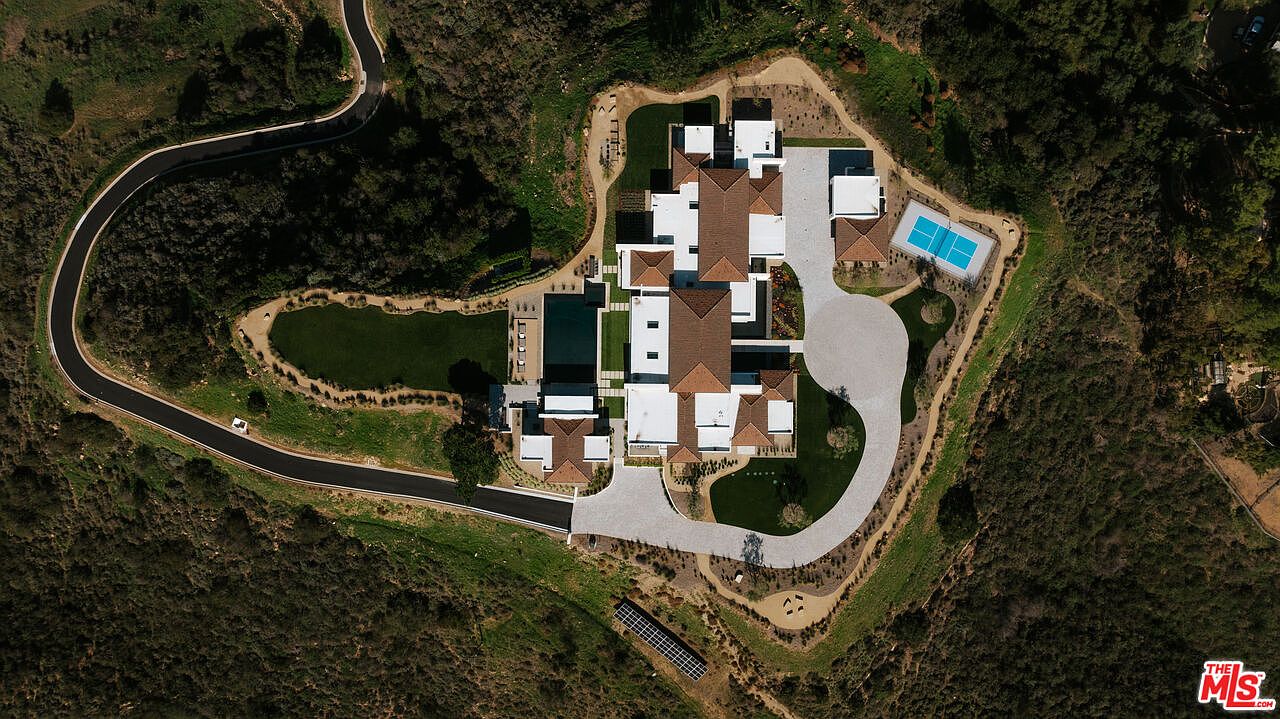 This high-altitude aerial view captures a sprawling luxury estate nestled within a secluded, hilly landscape. The property features a large multi-winged residence with a terracotta-tiled roof, a rectangular swimming pool, a private tennis court, and extensive manicured lawns connected by winding paved driveways. The perspective emphasizes the estate's privacy, grand scale, and seamless integration into the surrounding natural terrain.