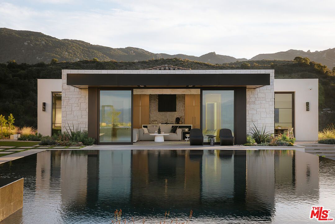 This stunning modern residence features a seamless indoor-outdoor transition, highlighted by a large infinity-edge pool that reflects the home's clean, contemporary architecture. The structure is characterized by stone masonry, expansive glass sliding doors, and a minimalist aesthetic, set against a backdrop of rolling hills. The overall impression is one of luxury, tranquility, and sophisticated design.