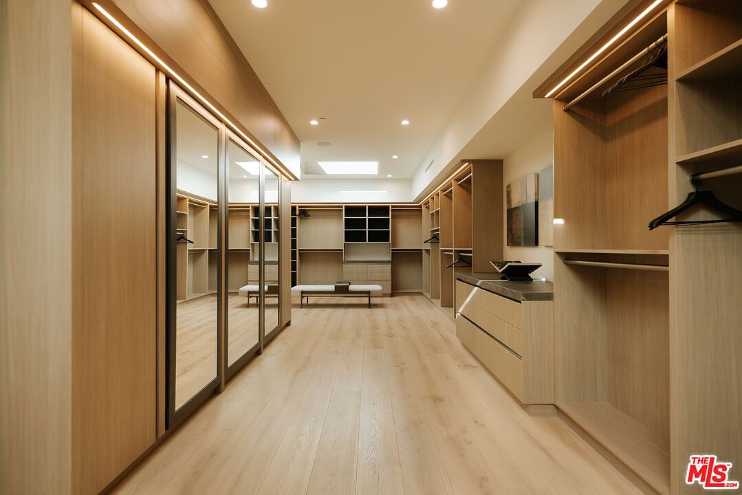 This expansive, high-end walk-in closet features light wood cabinetry, integrated LED lighting, and mirrored wardrobe doors that create a sense of depth and luxury. The room is anchored by a central bench and a sleek dresser island, offering a clean, minimalist aesthetic with ample storage space. The perspective is a centered, eye-level shot that emphasizes the symmetry and organized layout of this sophisticated dressing area.