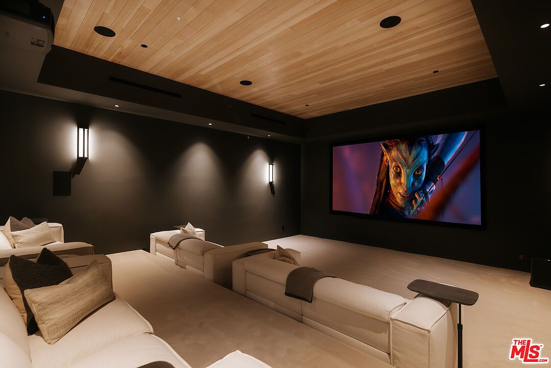 This image showcases a luxurious, modern home theater room designed for an immersive cinematic experience. The space features tiered seating with plush, light-colored sofas, a large projection screen displaying a vibrant movie scene, and a sophisticated wood-paneled ceiling with recessed lighting. The dark, moody wall color enhances the theater atmosphere, creating a private and high-end entertainment environment.