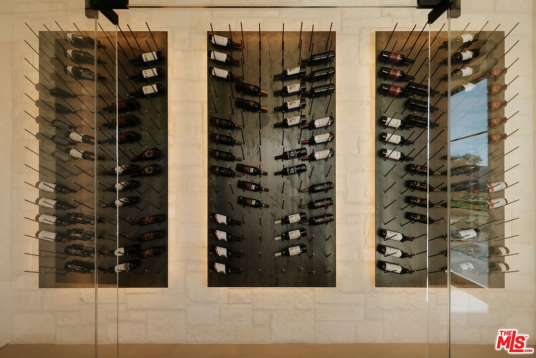 This sophisticated wine cellar features three vertical, glass-enclosed display racks set against a textured, light-colored stone wall. The minimalist metal racking system holds a curated collection of wine bottles, creating a sleek and modern aesthetic. The perspective is a direct, eye-level shot that emphasizes the symmetry and organized luxury of the storage space.