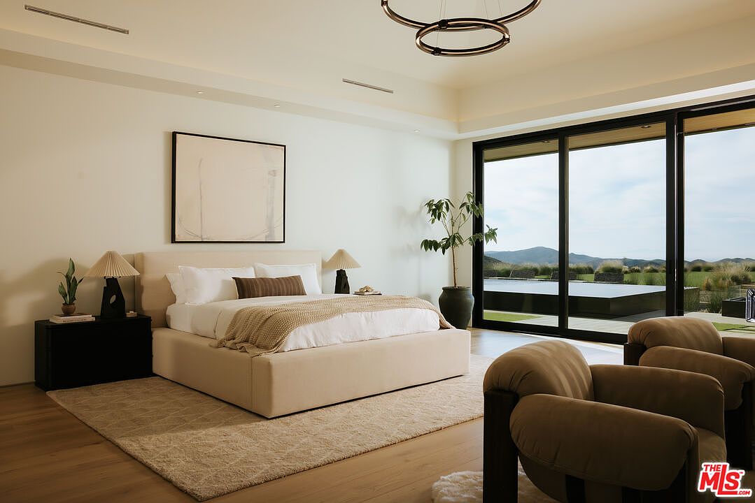 This luxurious primary bedroom features a minimalist, neutral aesthetic with a plush upholstered bed as the focal point, complemented by soft textures and warm wood flooring. Large floor-to-ceiling sliding glass doors provide an expansive view of the outdoor landscape and pool, seamlessly blending indoor comfort with the natural environment. The room is elegantly appointed with modern lighting fixtures and a potted plant, creating a serene and sophisticated atmosphere.
