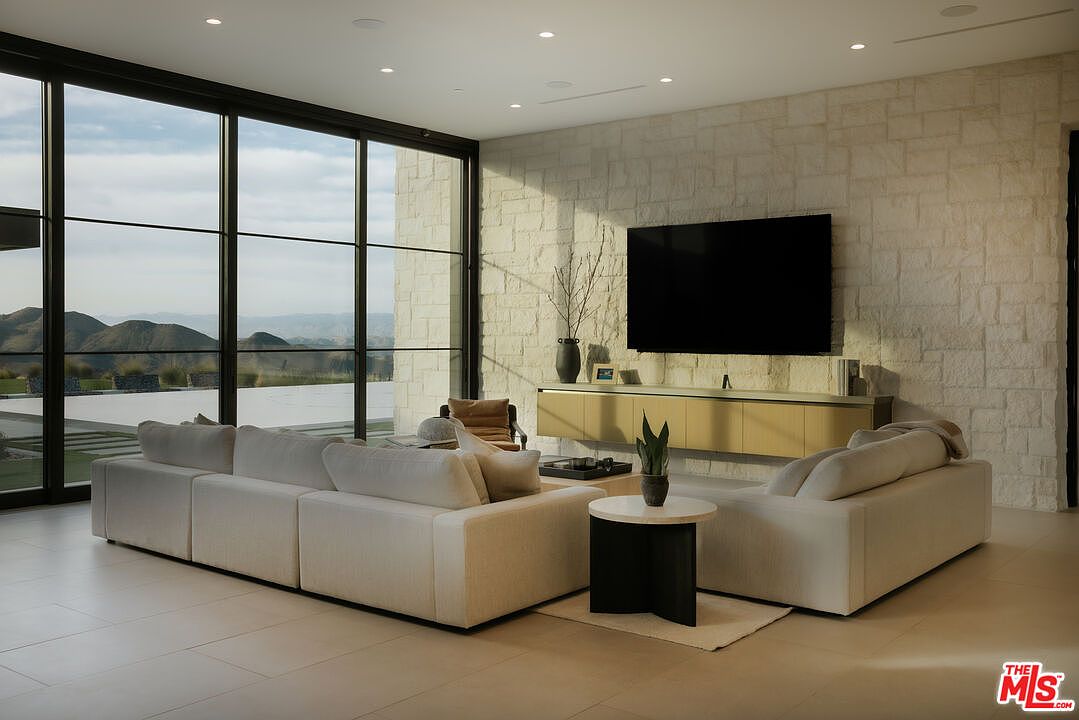 This sophisticated, modern living room features a large, plush white sectional sofa arranged around a minimalist circular side table, creating an inviting conversation area. A massive wall-mounted television sits above a sleek, floating gold-toned media console against a textured stone accent wall. Expansive floor-to-ceiling windows frame a breathtaking panoramic view of rolling hills and a serene outdoor pool, blending indoor luxury with the natural landscape.