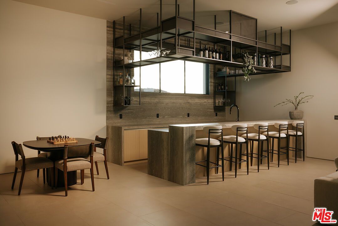 This sophisticated modern bar and lounge area features a striking stone-clad island with five sleek, upholstered bar stools. Above the counter, a custom industrial-style metal shelving unit provides elegant storage for glassware and bottles, while a nearby dining table set for chess adds a touch of refined leisure. The space is defined by its minimalist aesthetic, warm neutral tones, and high-end finishes, creating an inviting atmosphere for entertaining.
